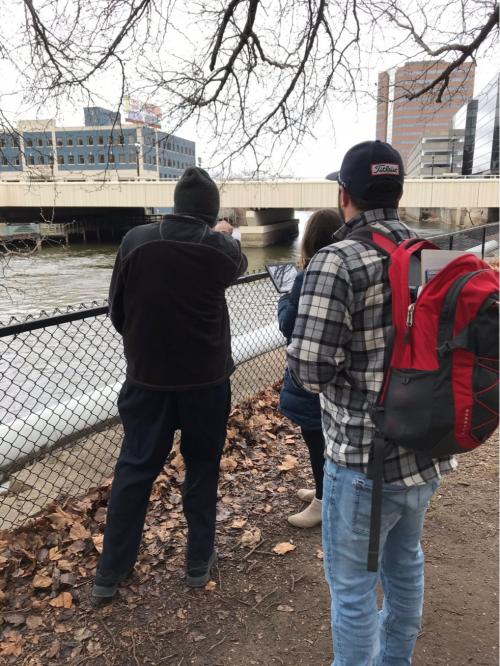 Three teachers stand next to the Grand River in winter, with city buildings in the background.
