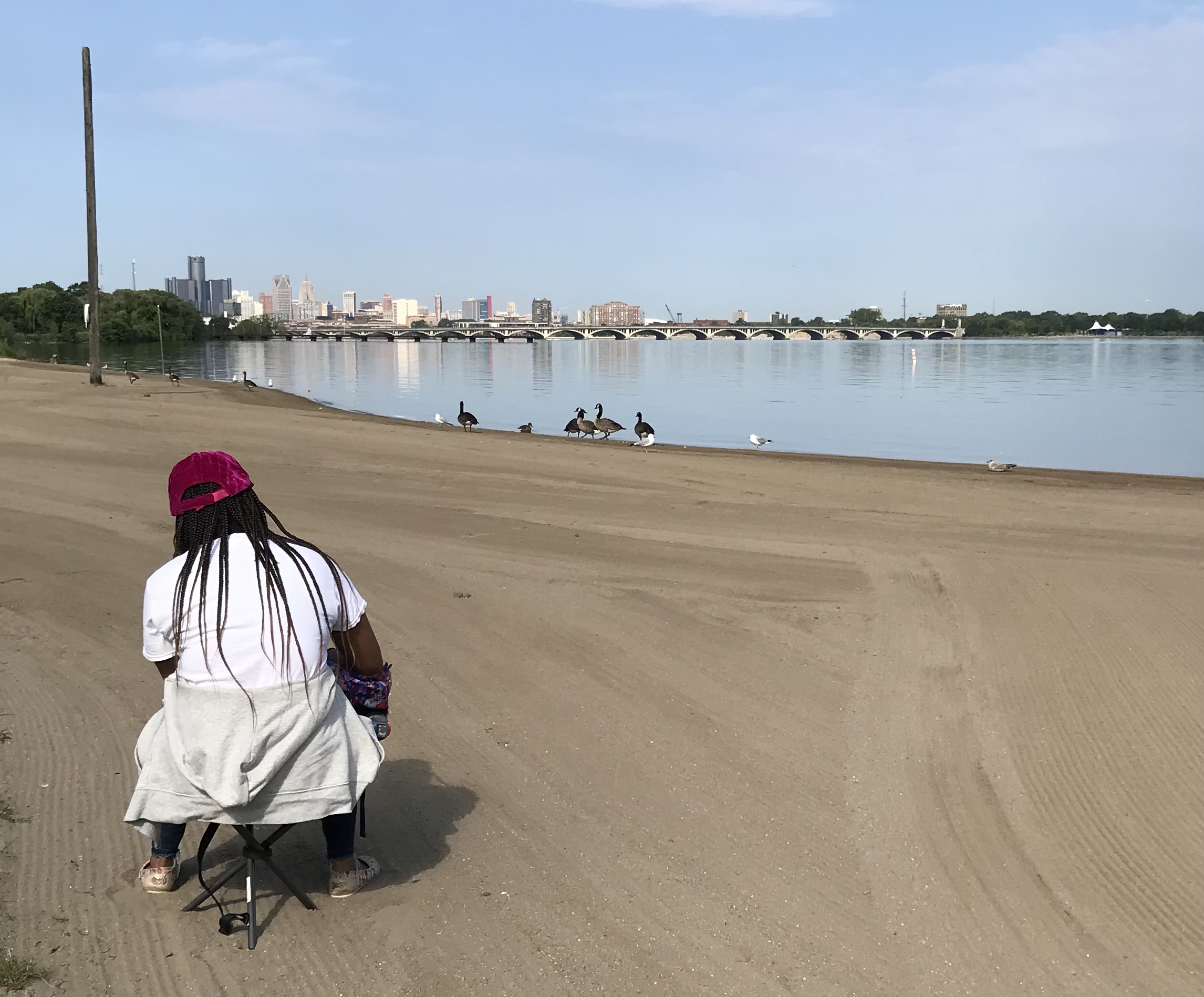 A teacher sitting on the beach observing birds with Detroit in the background